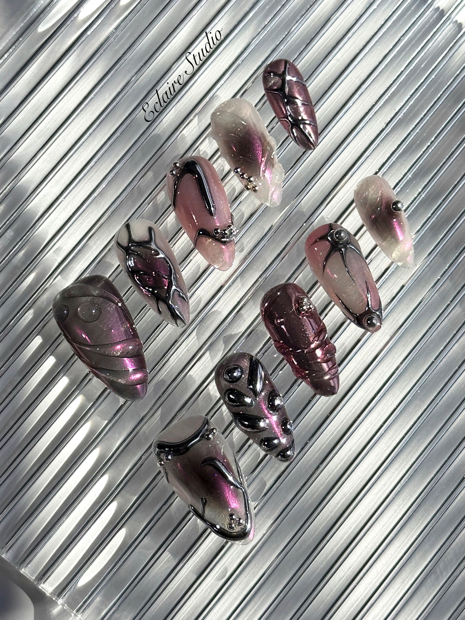 Set of long, almond-shaped press-on nails with a pink-to-purple multichrome base, accented by sculpted chrome designs, metallic textures, and small silver studs, displayed on a ribbed reflective surface under natural light.