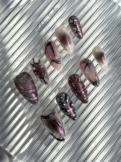 Set of long, almond-shaped press-on nails with a pink-to-purple multichrome base, accented by sculpted chrome designs, metallic textures, and small silver studs, displayed on a ribbed reflective surface under natural light.