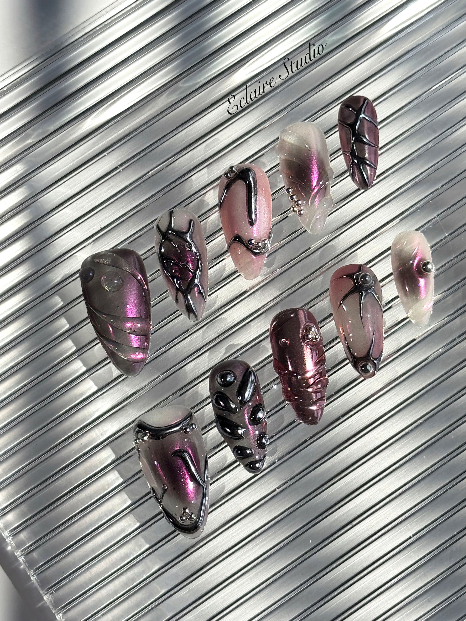 Set of long, almond-shaped press-on nails with a pink-to-purple multichrome base, accented by sculpted chrome designs, metallic textures, and small silver studs, displayed on a ribbed reflective surface under natural light.