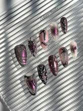 Set of long, almond-shaped press-on nails with a pink-to-purple multichrome base, accented by sculpted chrome designs, metallic textures, and small silver studs, displayed on a ribbed reflective surface under natural light.