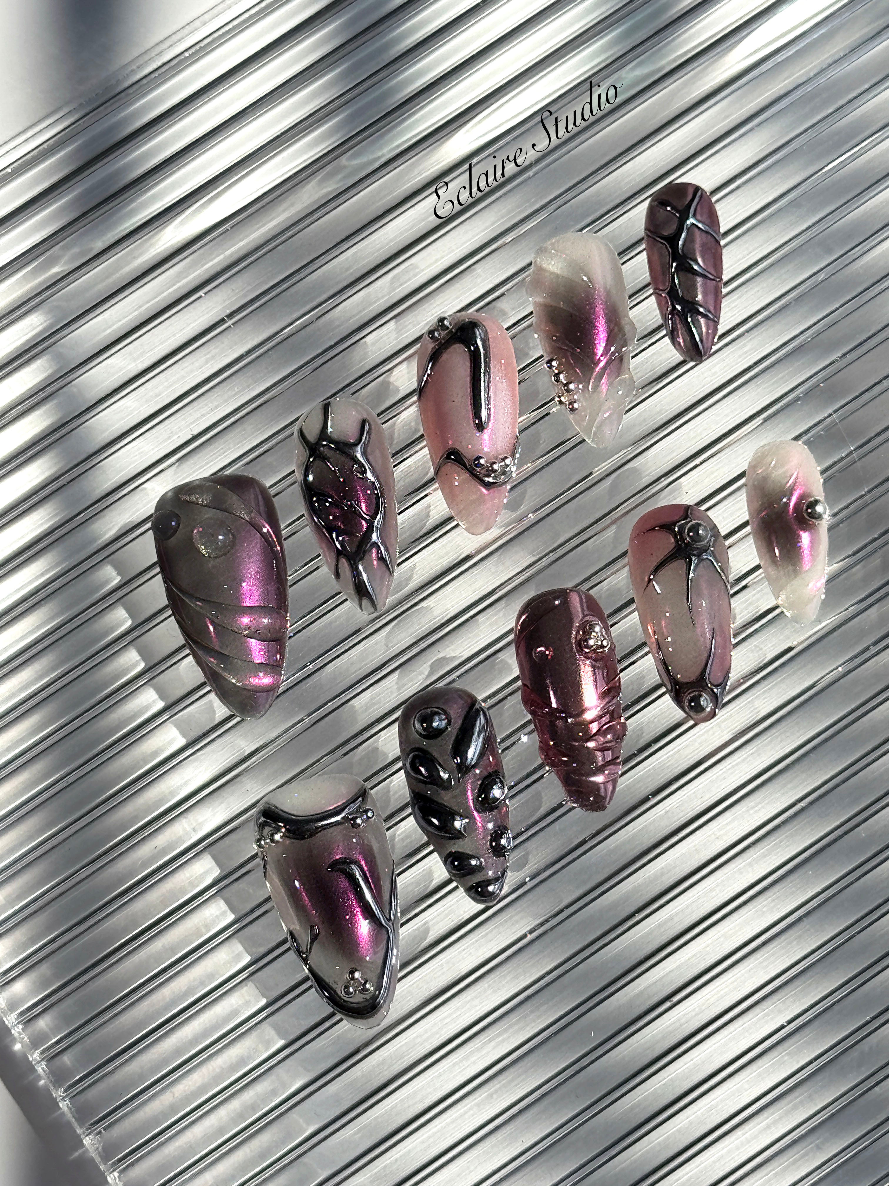 Set of long, almond-shaped press-on nails with a pink-to-purple multichrome base, accented by sculpted chrome designs, metallic textures, and small silver studs, displayed on a ribbed reflective surface under natural light.
