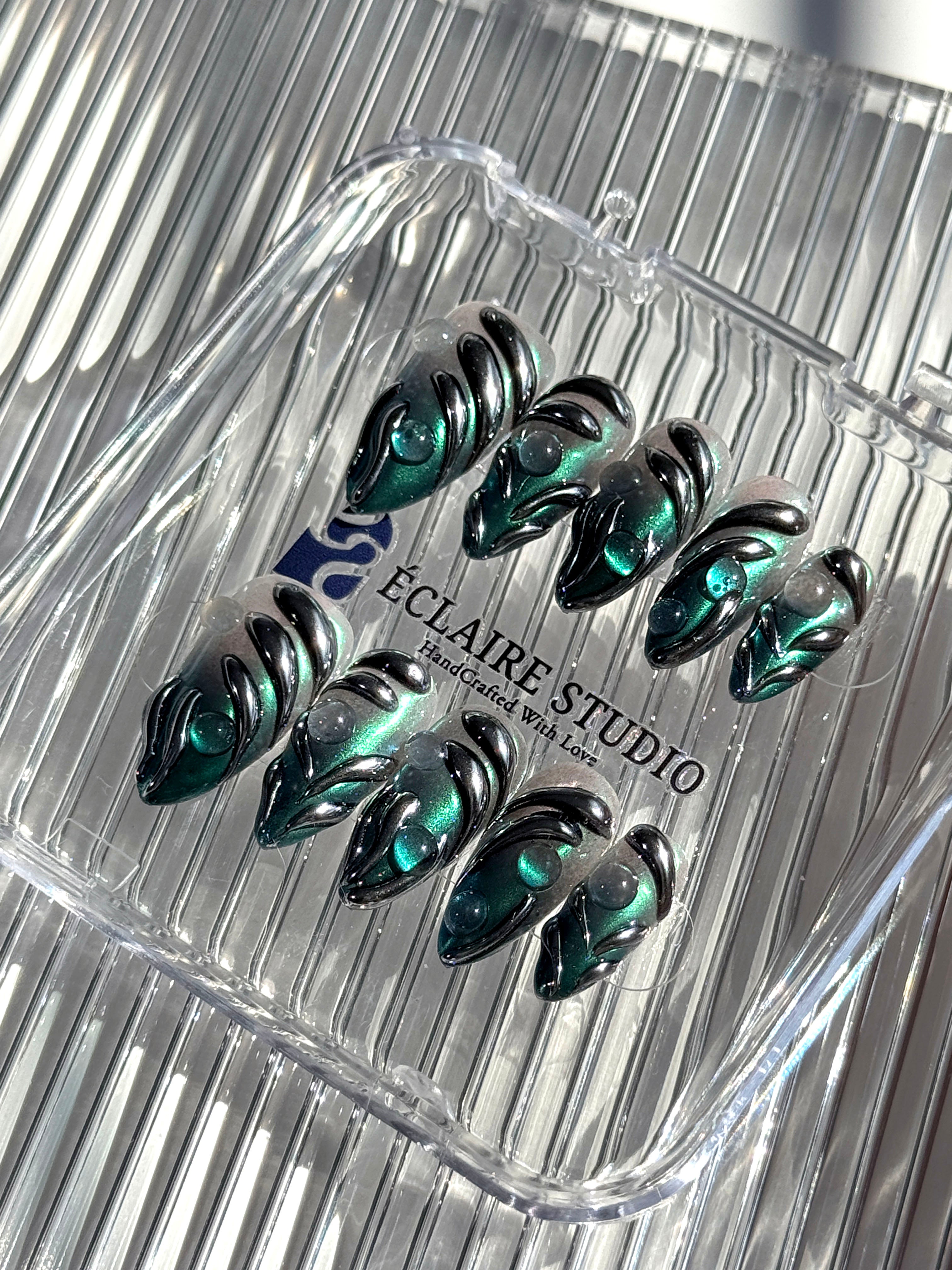Set of long almond-shaped press-on nails featuring a smoky green gradient base with metallic emerald green shimmer, accented by bold black chrome sculpted designs and glossy 3D details, displayed on a ribbed reflective surface under bright light.