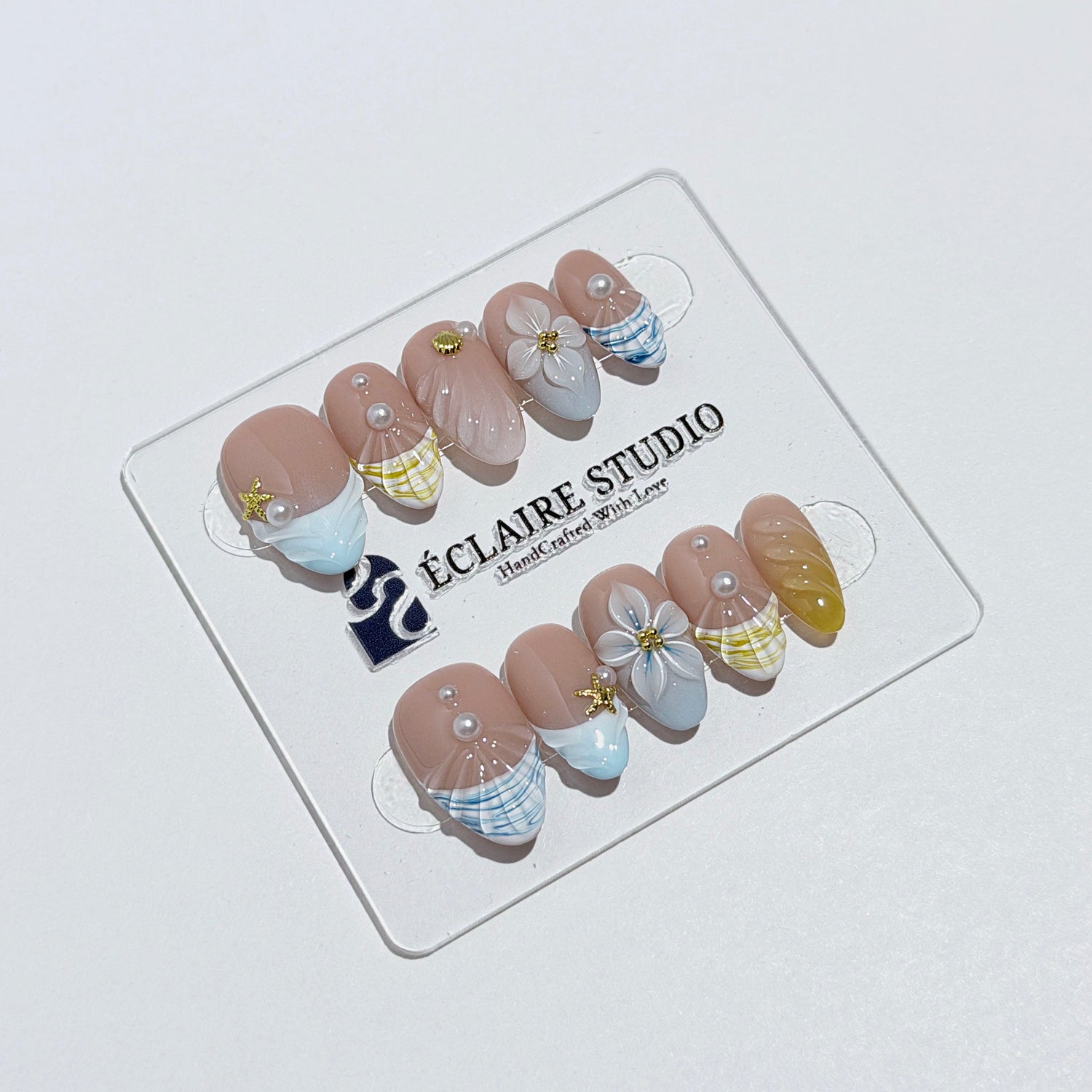 Artisanal resort-style press-on nails with a sheer nude base, pastel blue and yellow striped French tips, 3D sculpted flowers, and gold starfish charms with mini pearls.