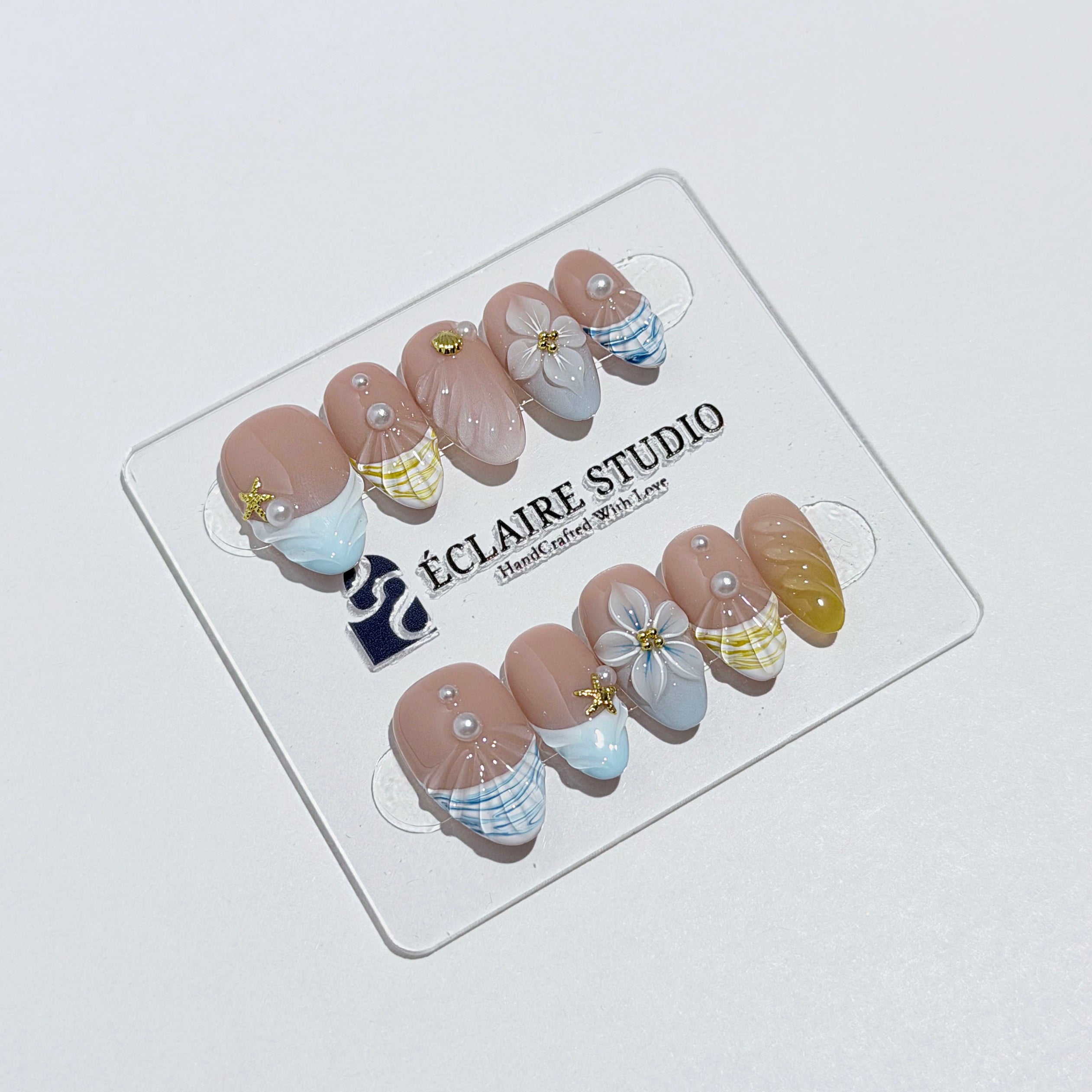 Artisanal resort-style press-on nails with a sheer nude base, pastel blue and yellow striped French tips, 3D sculpted flowers, and gold starfish charms with mini pearls.