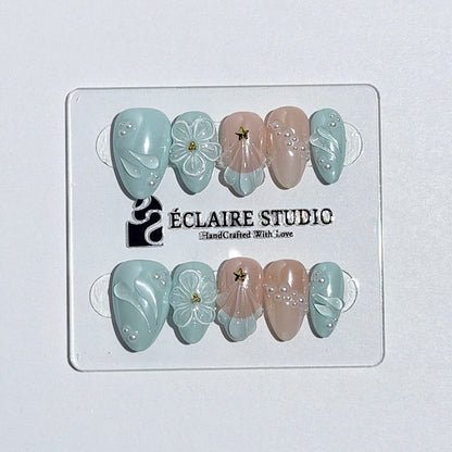 Eclaire Nail Studio 3D Summer short almond press-on nails featuring a light aqua glazed base and peachy-nude accent nails with 3D sculpted ocean waves, 3D flowers, and seashell tips with mini pearls and gold stars.