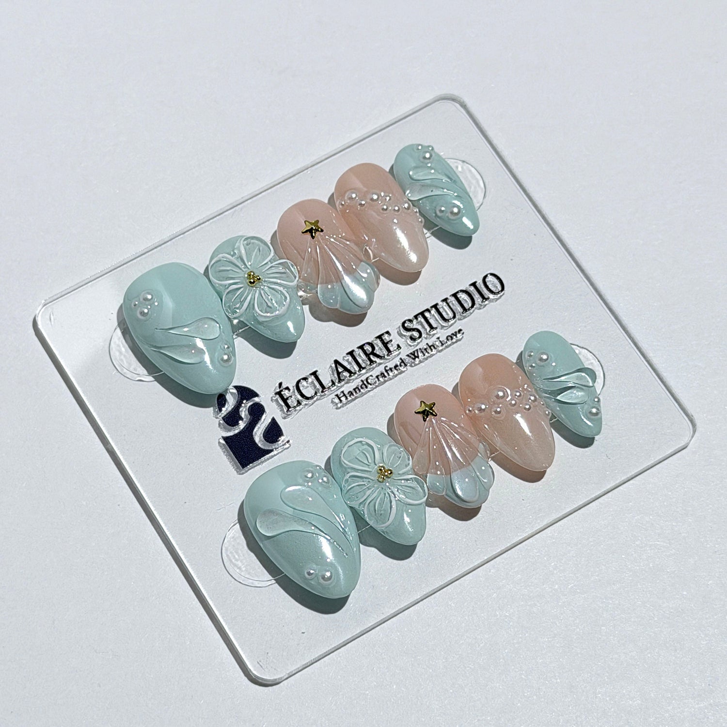 Eclaire Nail Studio 3D Summer short almond press-on nails featuring a light aqua glazed base and peachy-nude accent nails with 3D sculpted ocean waves, 3D flowers, and seashell tips with mini pearls and gold stars.