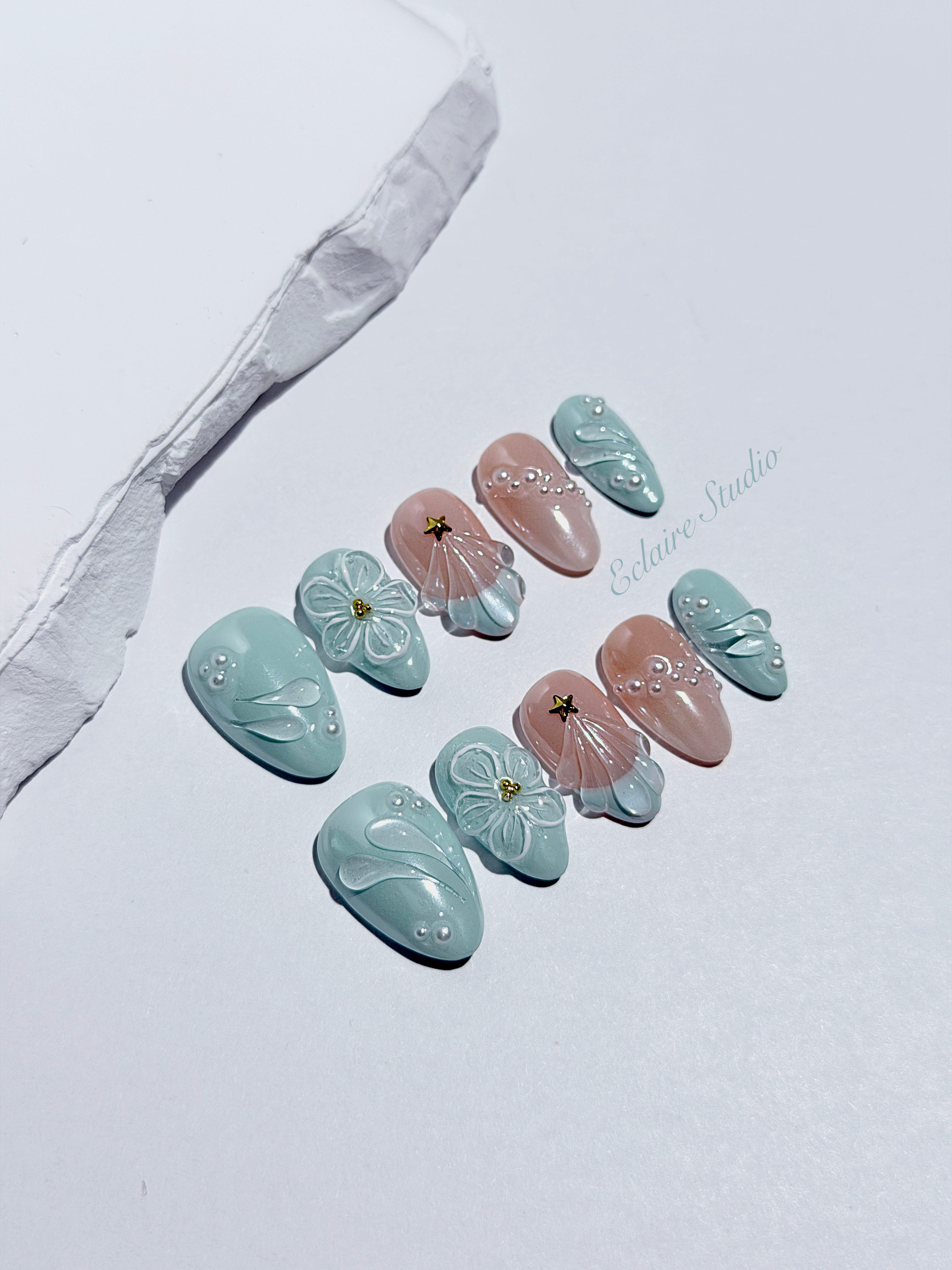 Eclaire Nail Studio 3D Summer short almond press-on nails featuring a light aqua glazed base and peachy-nude accent nails with 3D sculpted ocean waves, 3D flowers, and seashell tips with mini pearls and gold stars.