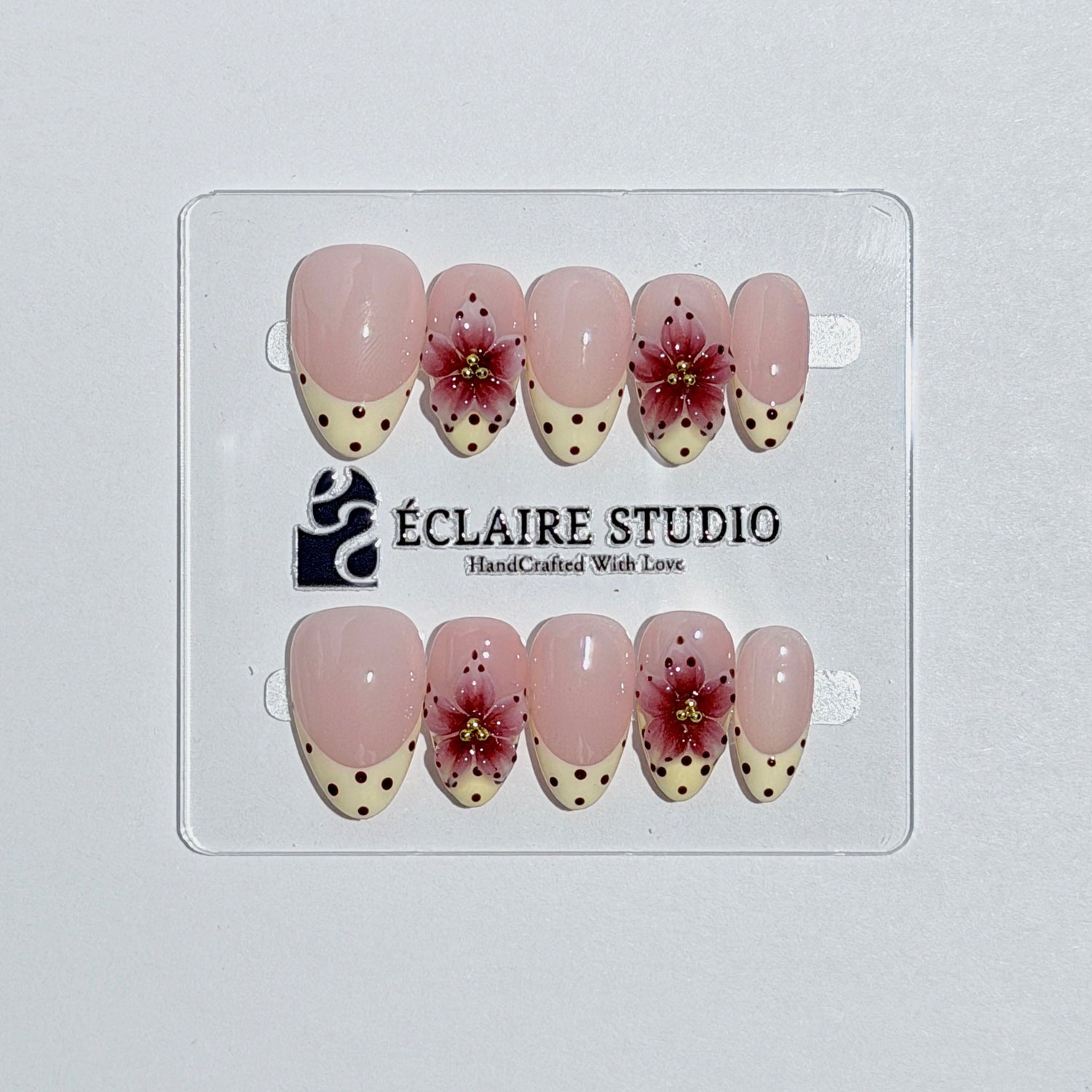 Luxury short almond press-on nails featuring sheer pink bases with vintage cream French tips, burgundy polka dots, and hand-sculpted 3D burgundy flowers with gold bead centers.