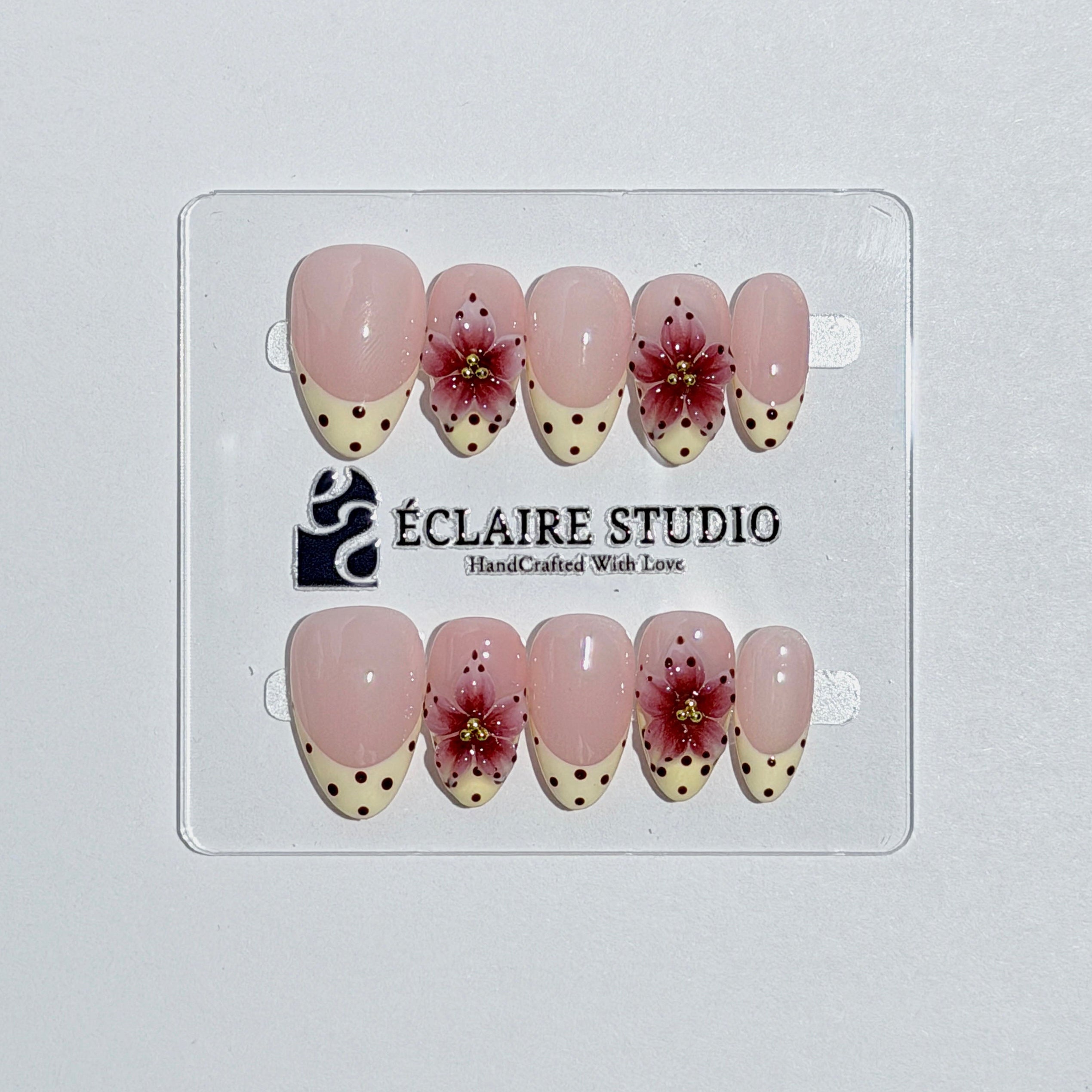 Luxury short almond press-on nails featuring sheer pink bases with vintage cream French tips, burgundy polka dots, and hand-sculpted 3D burgundy flowers with gold bead centers.