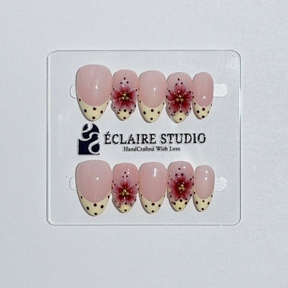 Luxury short almond press-on nails featuring sheer pink bases with vintage cream French tips, burgundy polka dots, and hand-sculpted 3D burgundy flowers with gold bead centers.