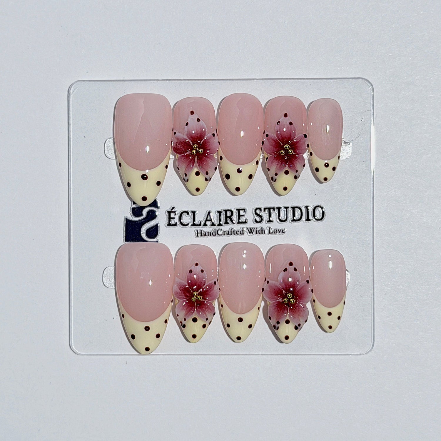 Eclaire Nail Studio Luxury medium almond press-on nails featuring sheer pink bases with vintage cream French tips, burgundy polka dots, and hand-sculpted 3D burgundy flowers with gold bead centers.
