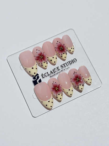 Luxury medium almond press-on nails featuring sheer pink bases with vintage cream French tips, burgundy polka dots, and hand-sculpted 3D burgundy flowers with gold bead centers.