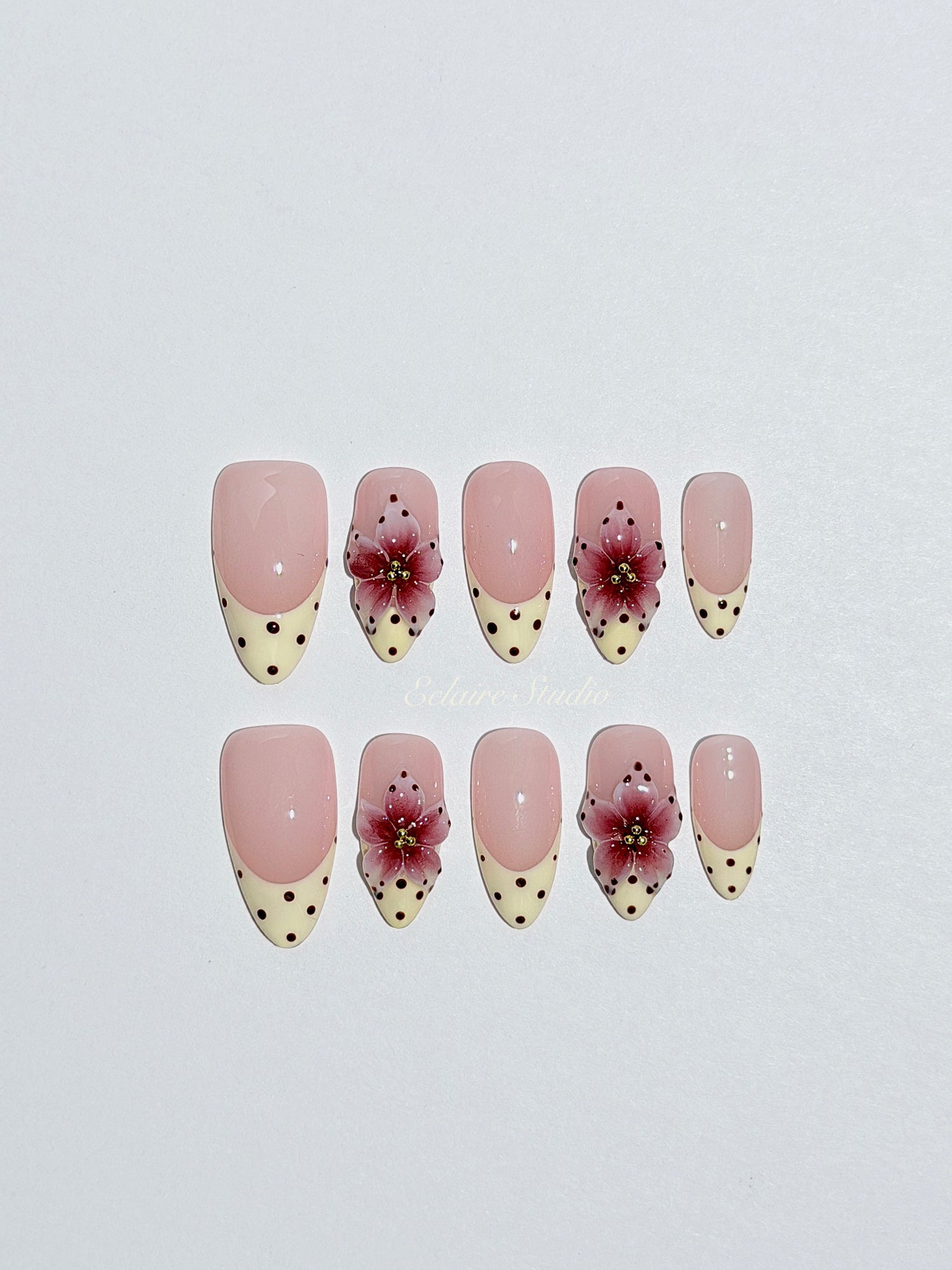 Luxury medium almond press-on nails featuring sheer pink bases with vintage cream French tips, burgundy polka dots, and hand-sculpted 3D burgundy flowers with gold bead centers.