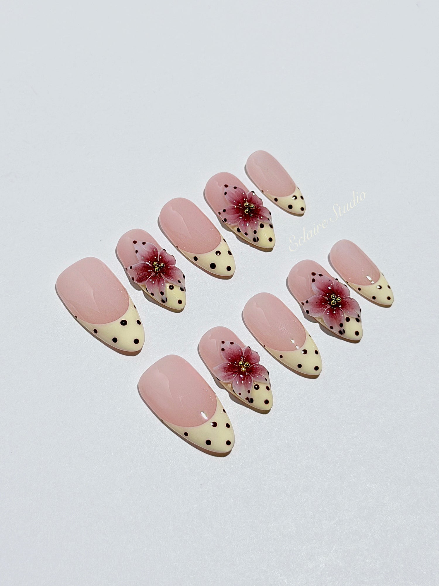 Luxury medium almond press-on nails featuring sheer pink bases with vintage cream French tips, burgundy polka dots, and hand-sculpted 3D burgundy flowers with gold bead centers.