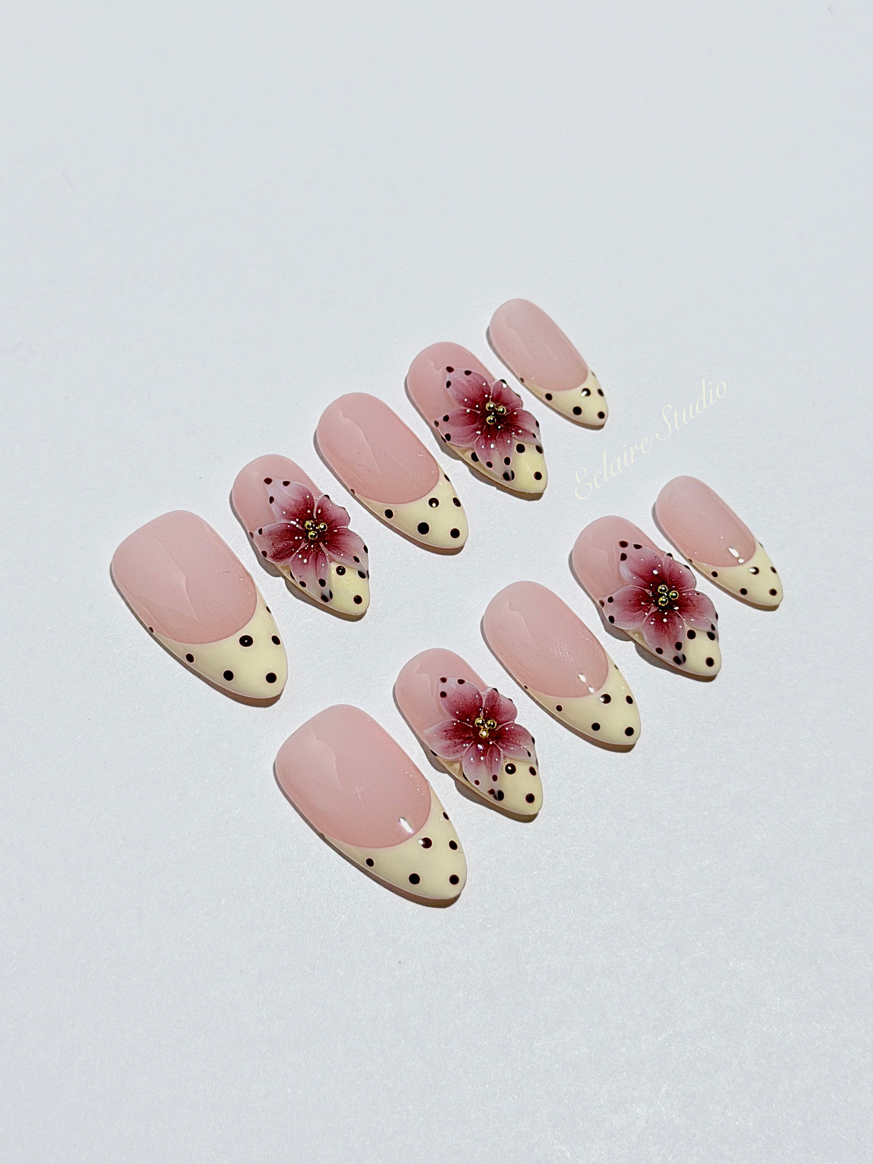 Luxury medium almond press-on nails featuring sheer pink bases with vintage cream French tips, burgundy polka dots, and hand-sculpted 3D burgundy flowers with gold bead centers.