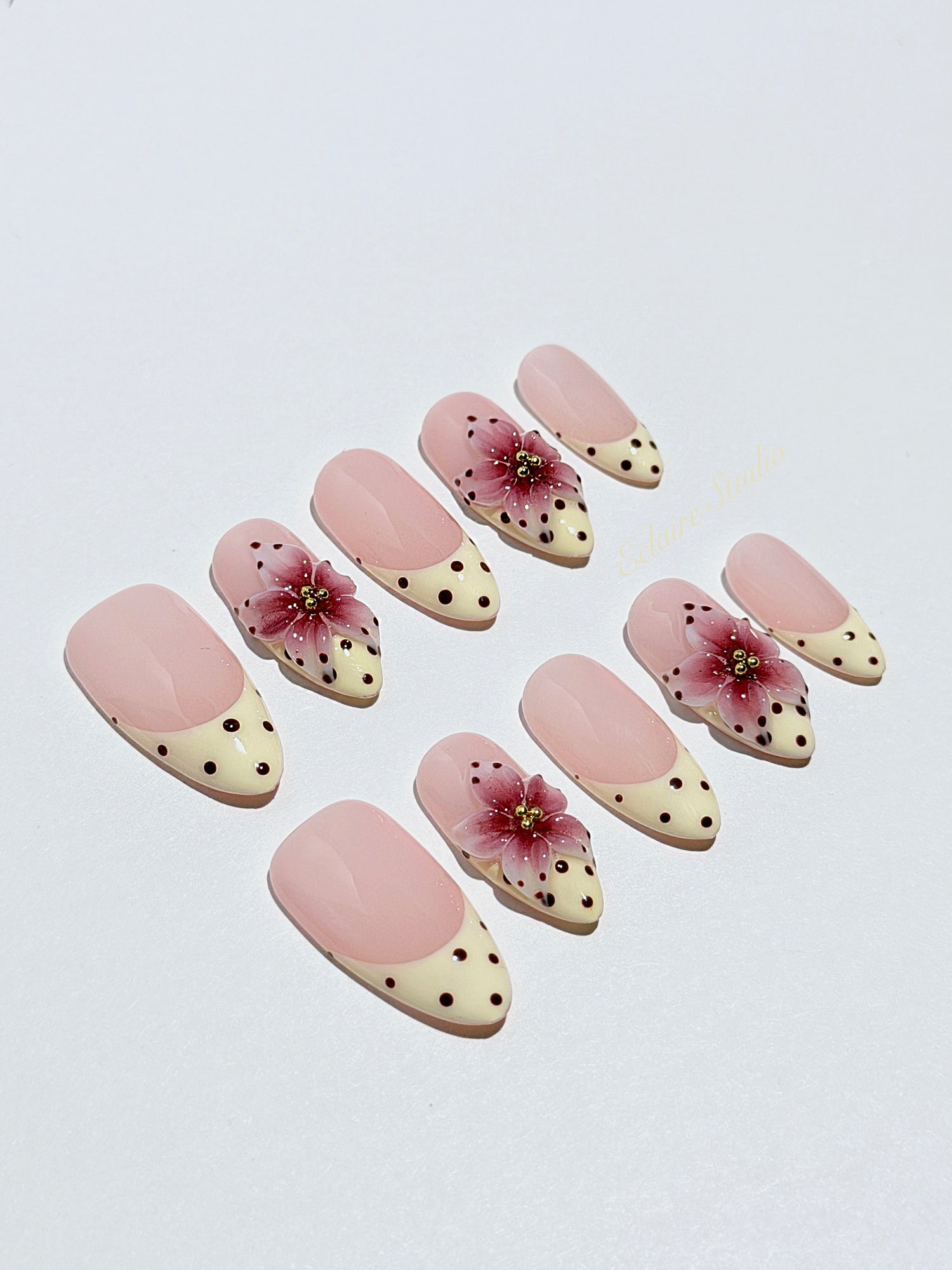 Luxury medium almond press-on nails featuring sheer pink bases with vintage cream French tips, burgundy polka dots, and hand-sculpted 3D burgundy flowers with gold bead centers.