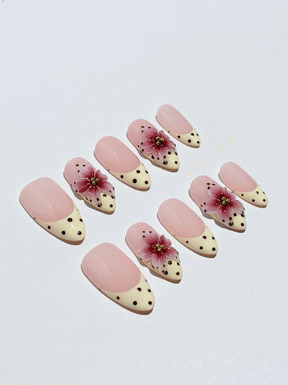 Luxury medium almond press-on nails featuring sheer pink bases with vintage cream French tips, burgundy polka dots, and hand-sculpted 3D burgundy flowers with gold bead centers.
