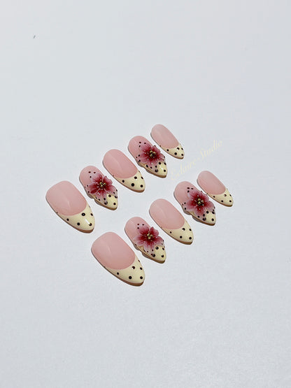 Luxury medium almond press-on nails featuring sheer pink bases with vintage cream French tips, burgundy polka dots, and hand-sculpted 3D burgundy flowers with gold bead centers.