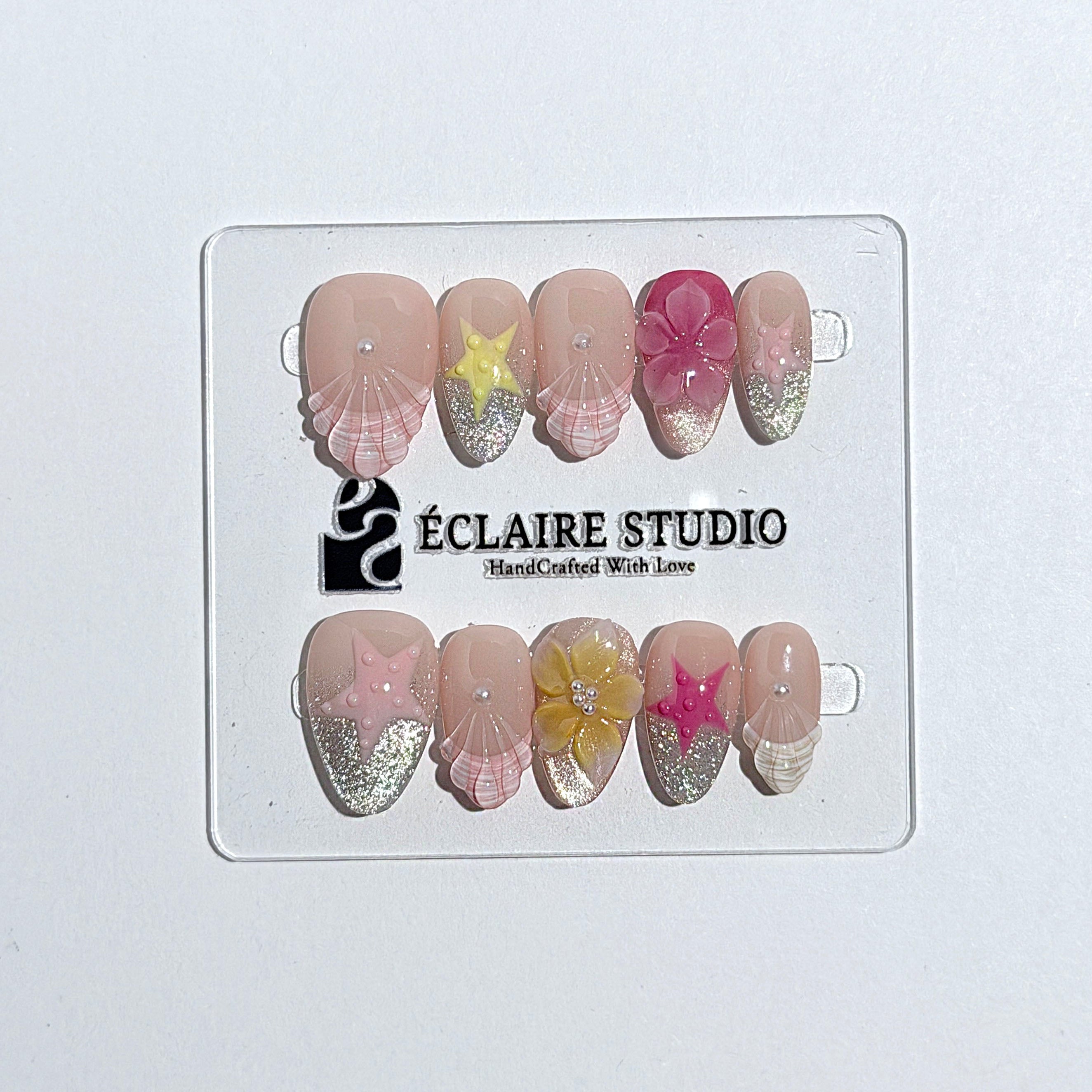 Eclaire Studio 3D Summer short almond press-on nails with sheer nude bases, sparkling silver cat-eye magnetic French tips, hand-painted colorful starfish, and pink 3D sculpted seashells.