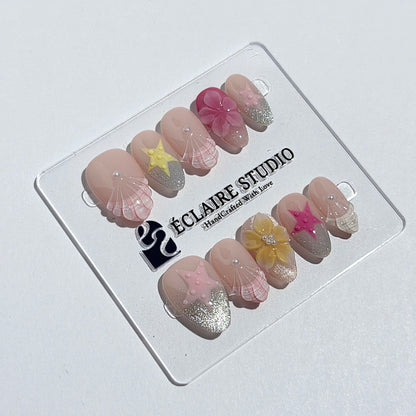 Eclaire Studio 3D Summer short almond press-on nails with sheer nude bases, sparkling silver cat-eye magnetic French tips, hand-painted colorful starfish, and pink 3D sculpted seashells.