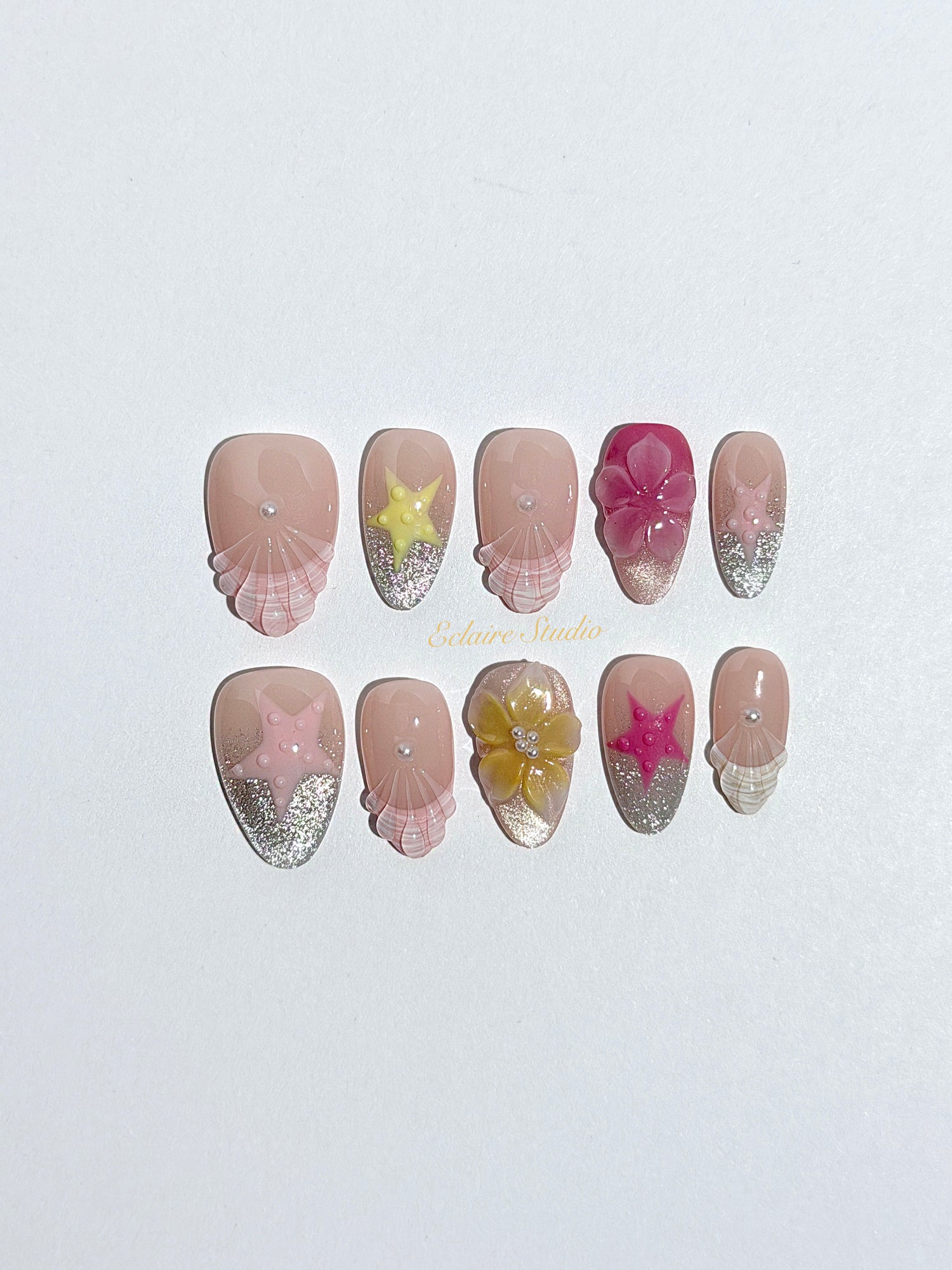 Eclaire Studio 3D Summer short almond press-on nails with sheer nude bases, sparkling silver cat-eye magnetic French tips, hand-painted colorful starfish, and pink 3D sculpted seashells.