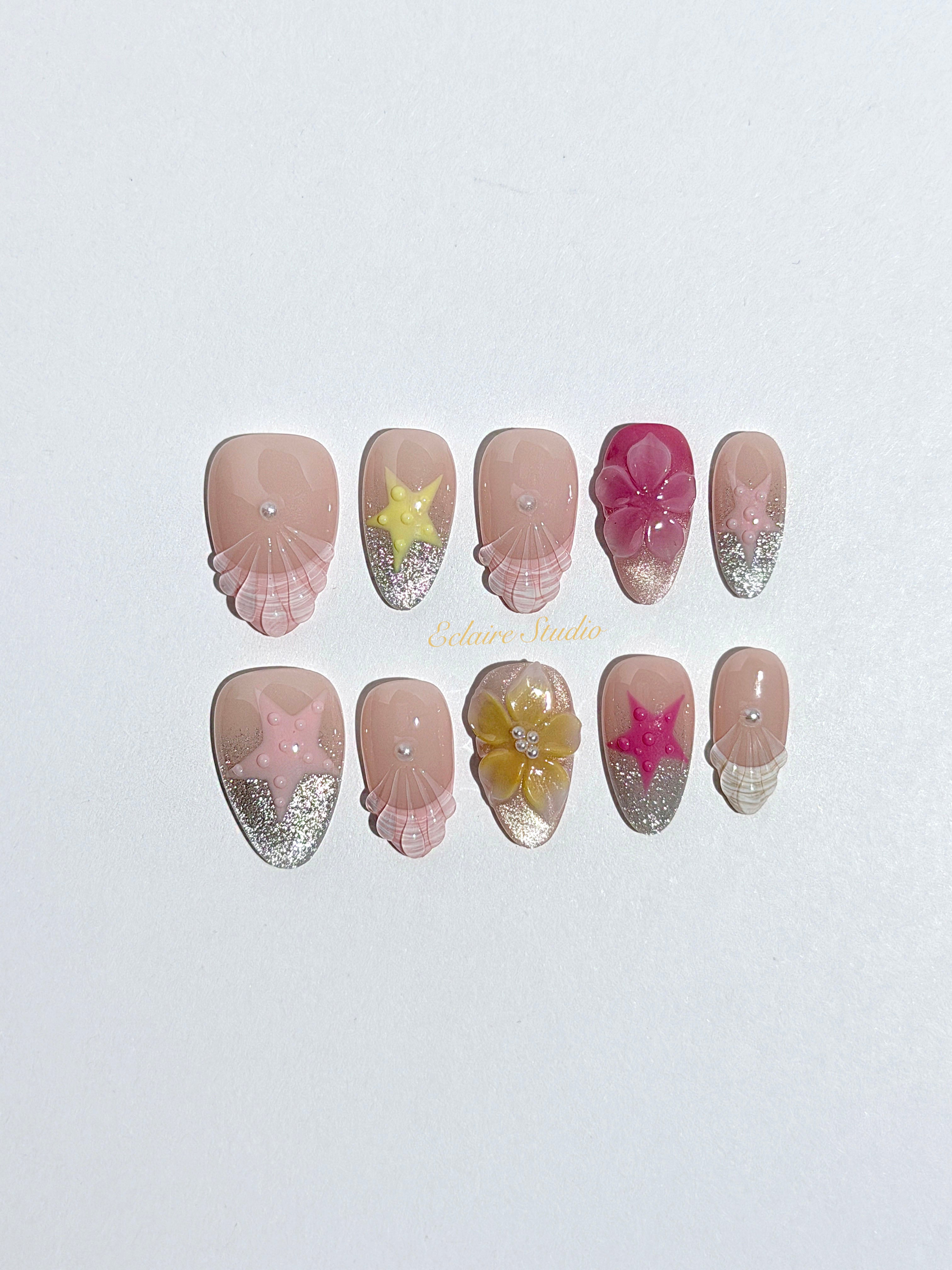 Eclaire Studio 3D Summer short almond press-on nails with sheer nude bases, sparkling silver cat-eye magnetic French tips, hand-painted colorful starfish, and pink 3D sculpted seashells.
