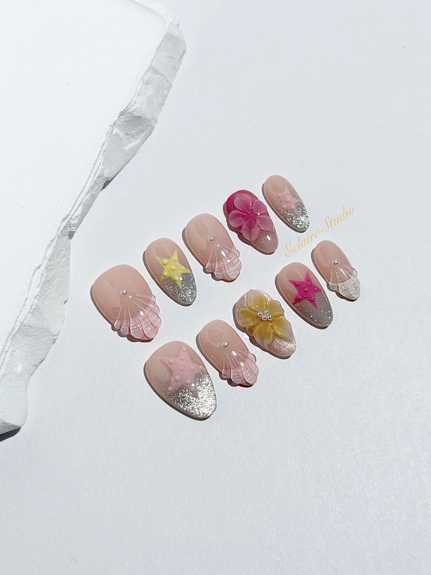 Eclaire Studio 3D Summer short almond press-on nails with sheer nude bases, sparkling silver cat-eye magnetic French tips, hand-painted colorful starfish, and pink 3D sculpted seashells.