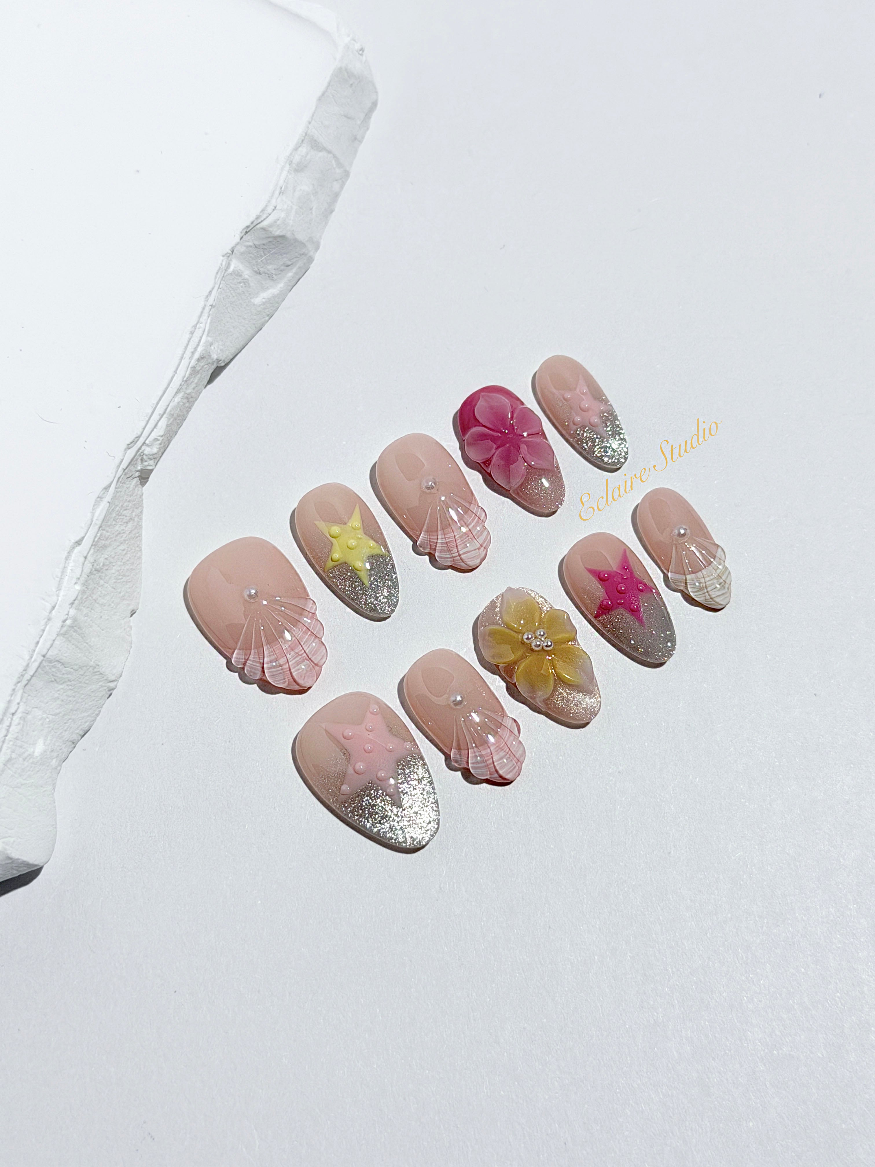 Eclaire Studio 3D Summer short almond press-on nails with sheer nude bases, sparkling silver cat-eye magnetic French tips, hand-painted colorful starfish, and pink 3D sculpted seashells.
