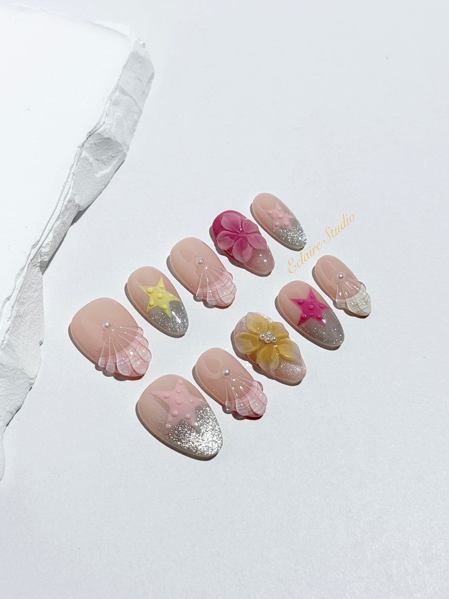 Eclaire Studio 3D Summer short almond press-on nails with sheer nude bases, sparkling silver cat-eye magnetic French tips, hand-painted colorful starfish, and pink 3D sculpted seashells.