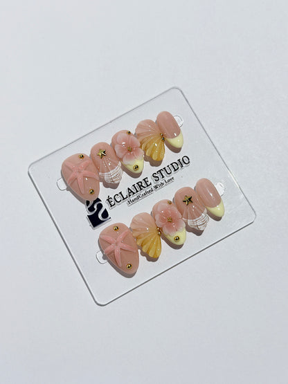 Summer Eclaire Nail Studio Luxury short almond press-on nails featuring a sheer nude base with butter yellow French tips, hand-sculpted 3D pink starfish, amber-tinted 3D seashells, and flowers with gold accents.