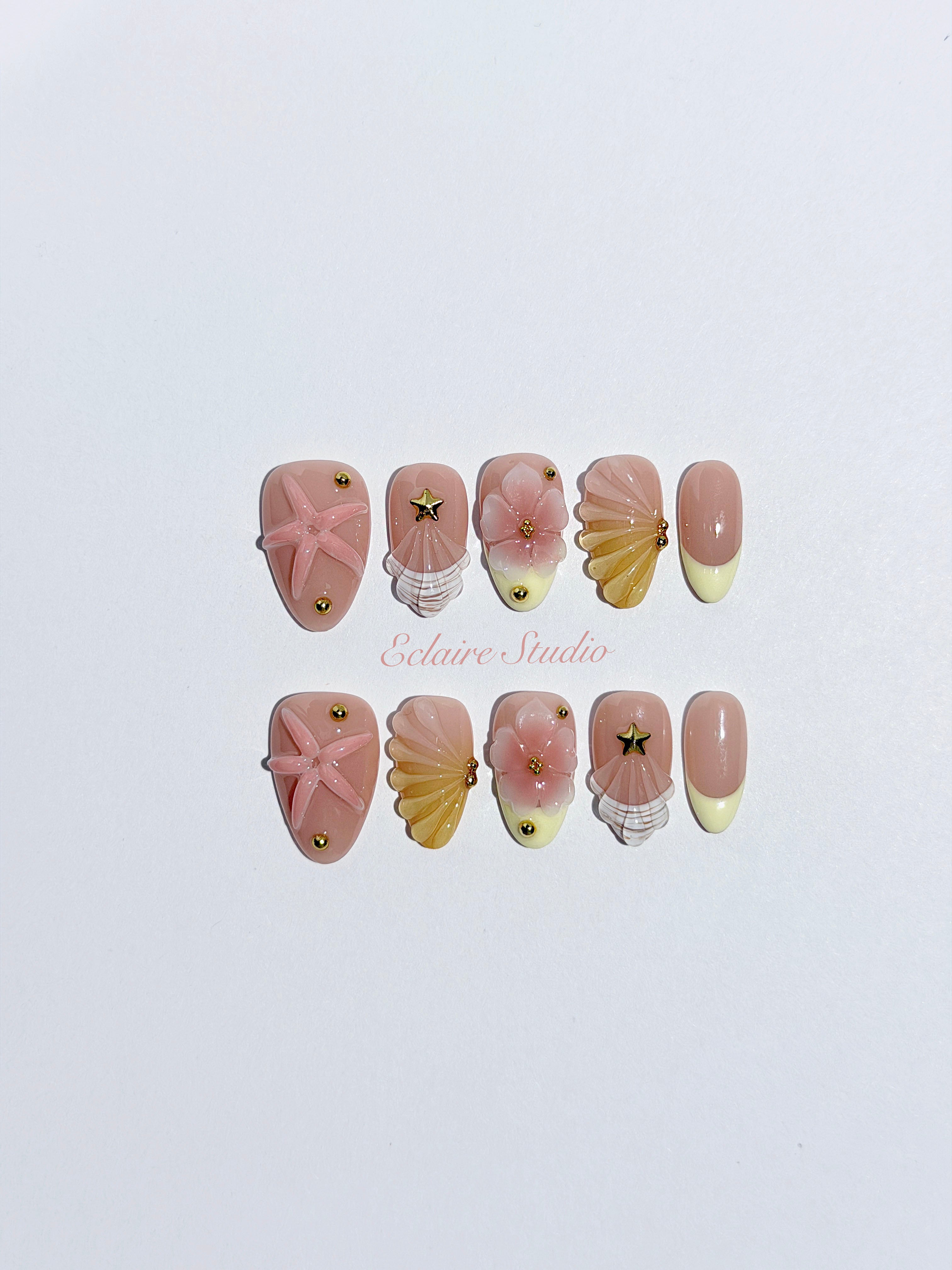 Summer Eclaire Nail Studio Luxury short almond press-on nails featuring a sheer nude base with butter yellow French tips, hand-sculpted 3D pink starfish, amber-tinted 3D seashells, and flowers with gold accents.