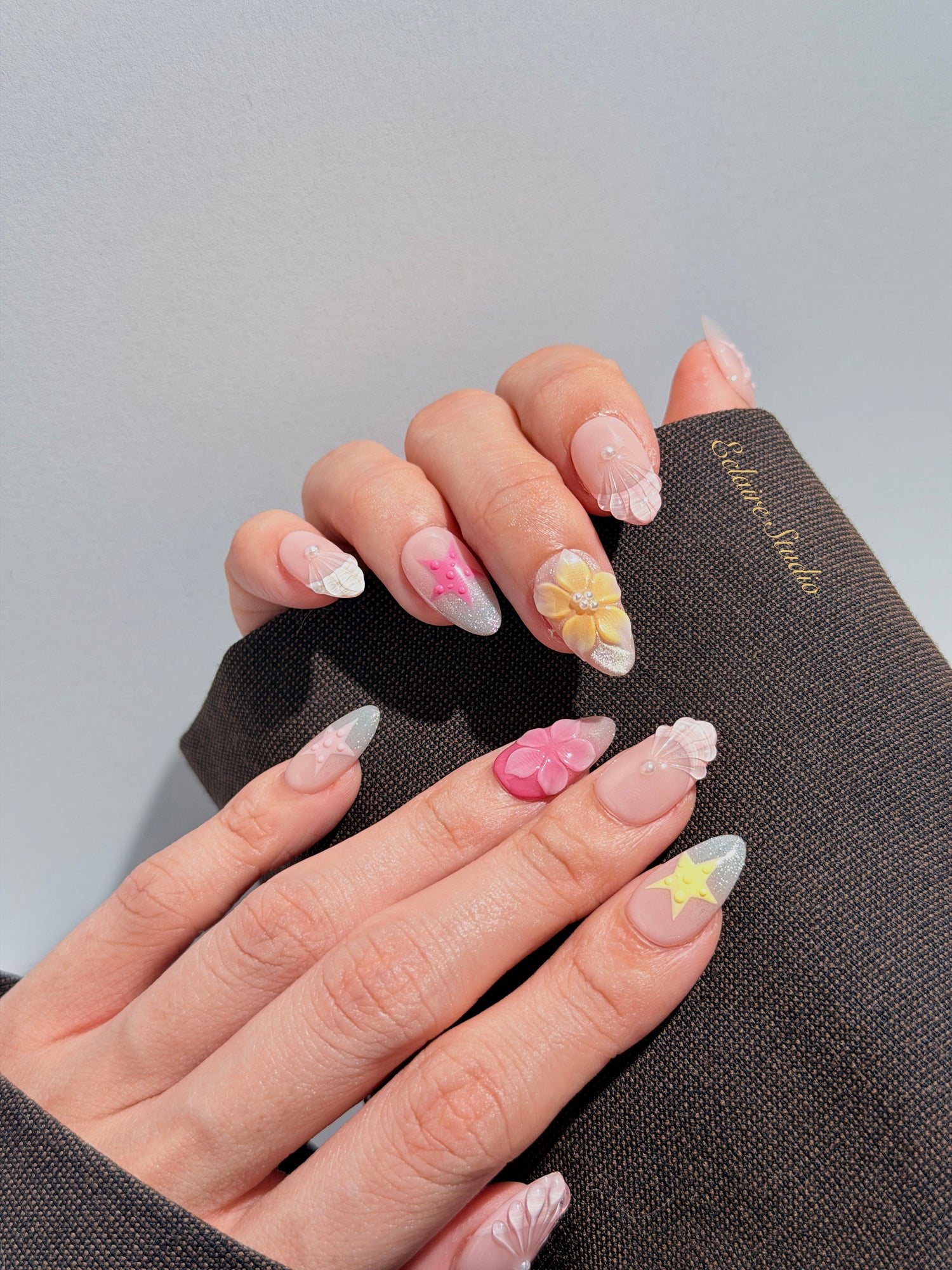 Eclaire Studio 3D Summer short almond press-on nails with sheer nude bases, sparkling silver cat-eye magnetic French tips, hand-painted colorful starfish, and pink 3D sculpted seashells.