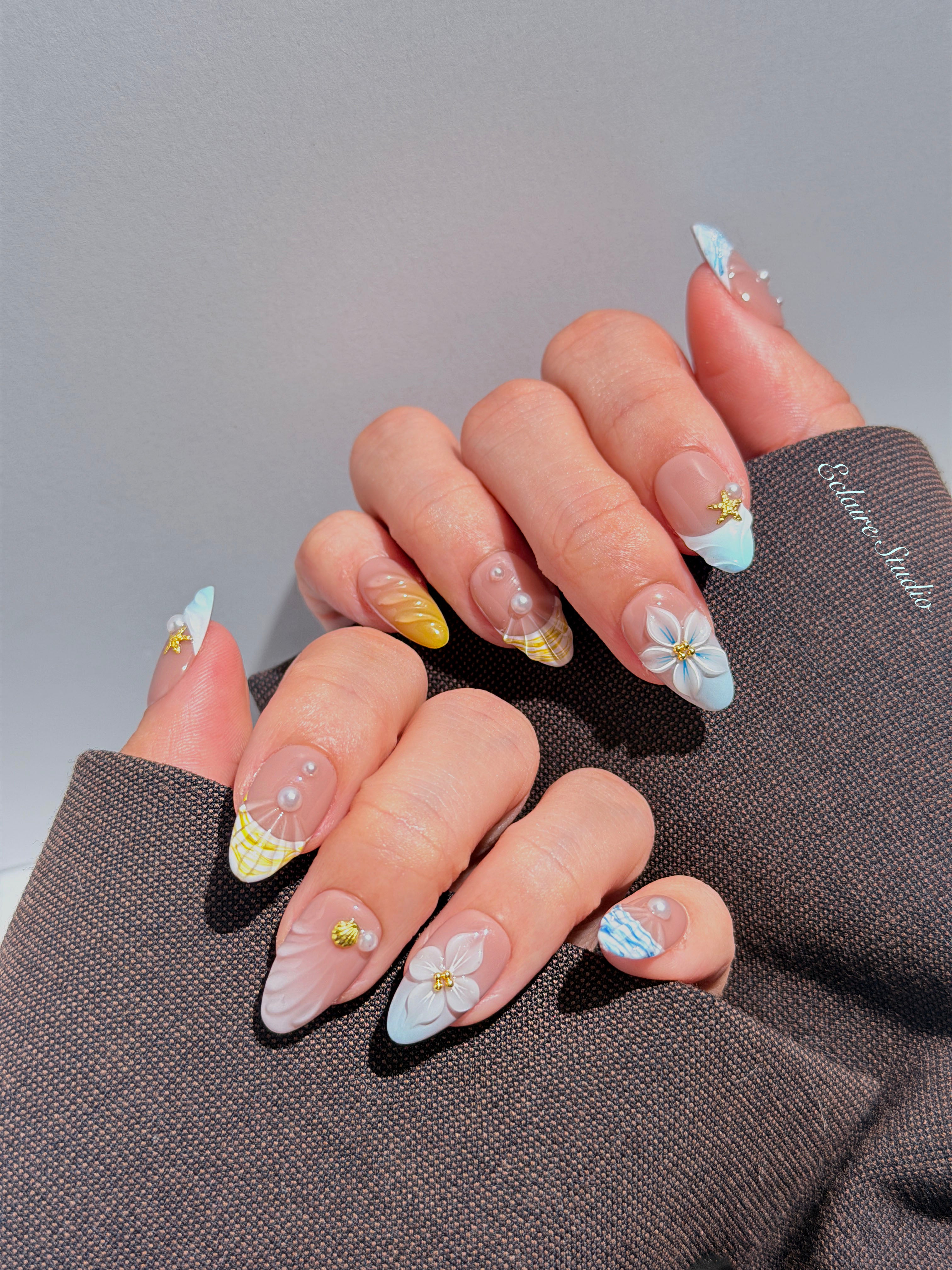 Artisanal resort-style press-on nails with a sheer nude base, pastel blue and yellow striped French tips, 3D sculpted flowers, and gold starfish charms with mini pearls.