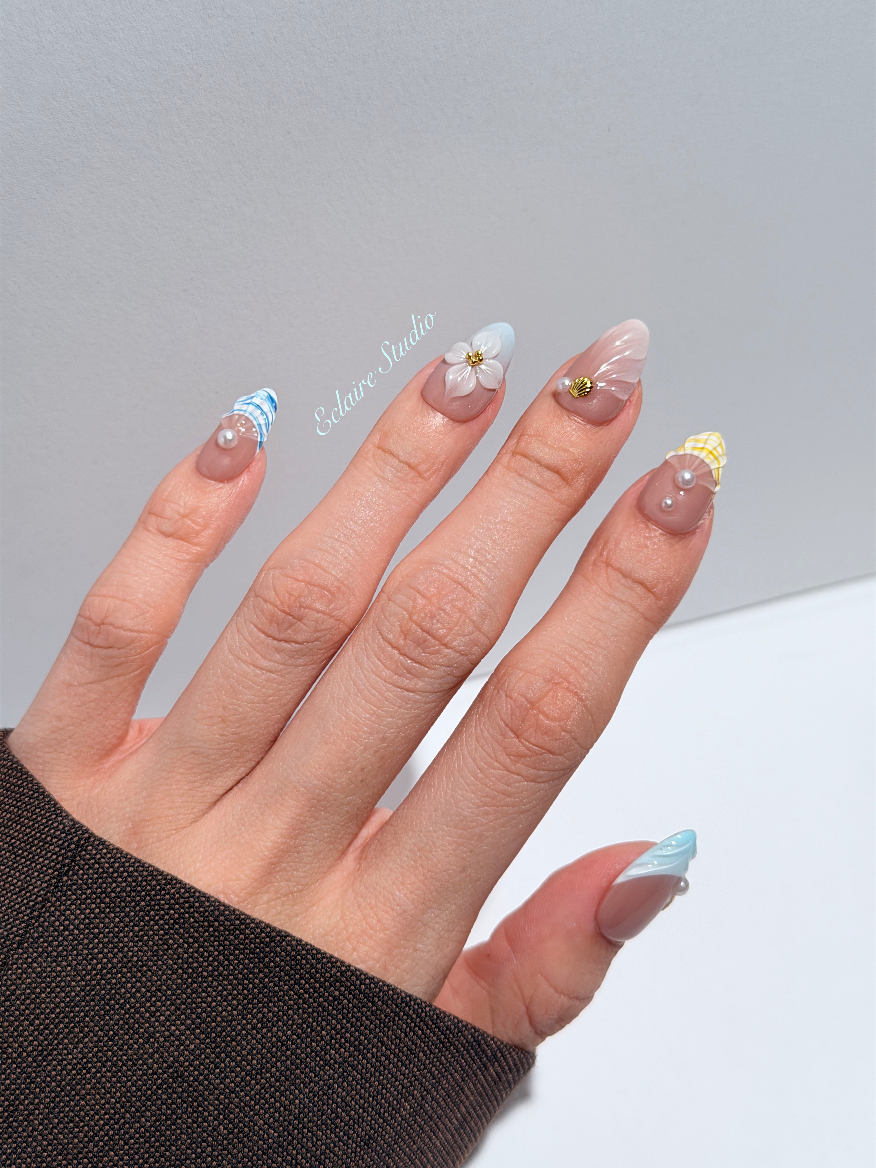 Artisanal resort-style press-on nails with a sheer nude base, pastel blue and yellow striped French tips, 3D sculpted flowers, and gold starfish charms with mini pearls.
