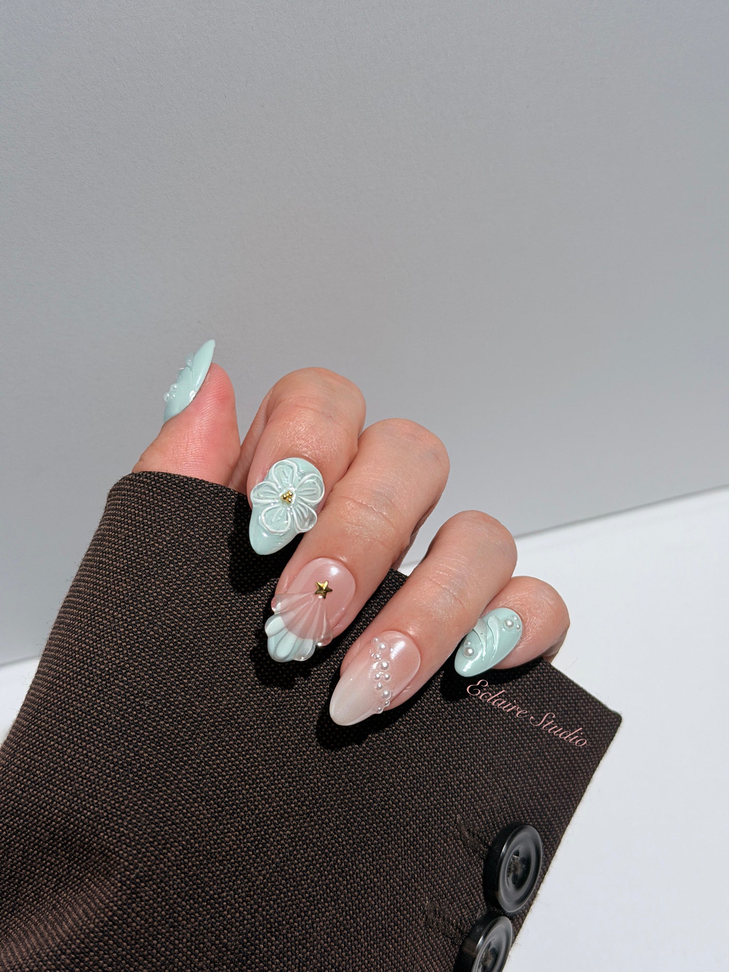 Eclaire Nail Studio 3D Summer short almond press-on nails featuring a light aqua glazed base and peachy-nude accent nails with 3D sculpted ocean waves, 3D flowers, and seashell tips with mini pearls and gold stars.