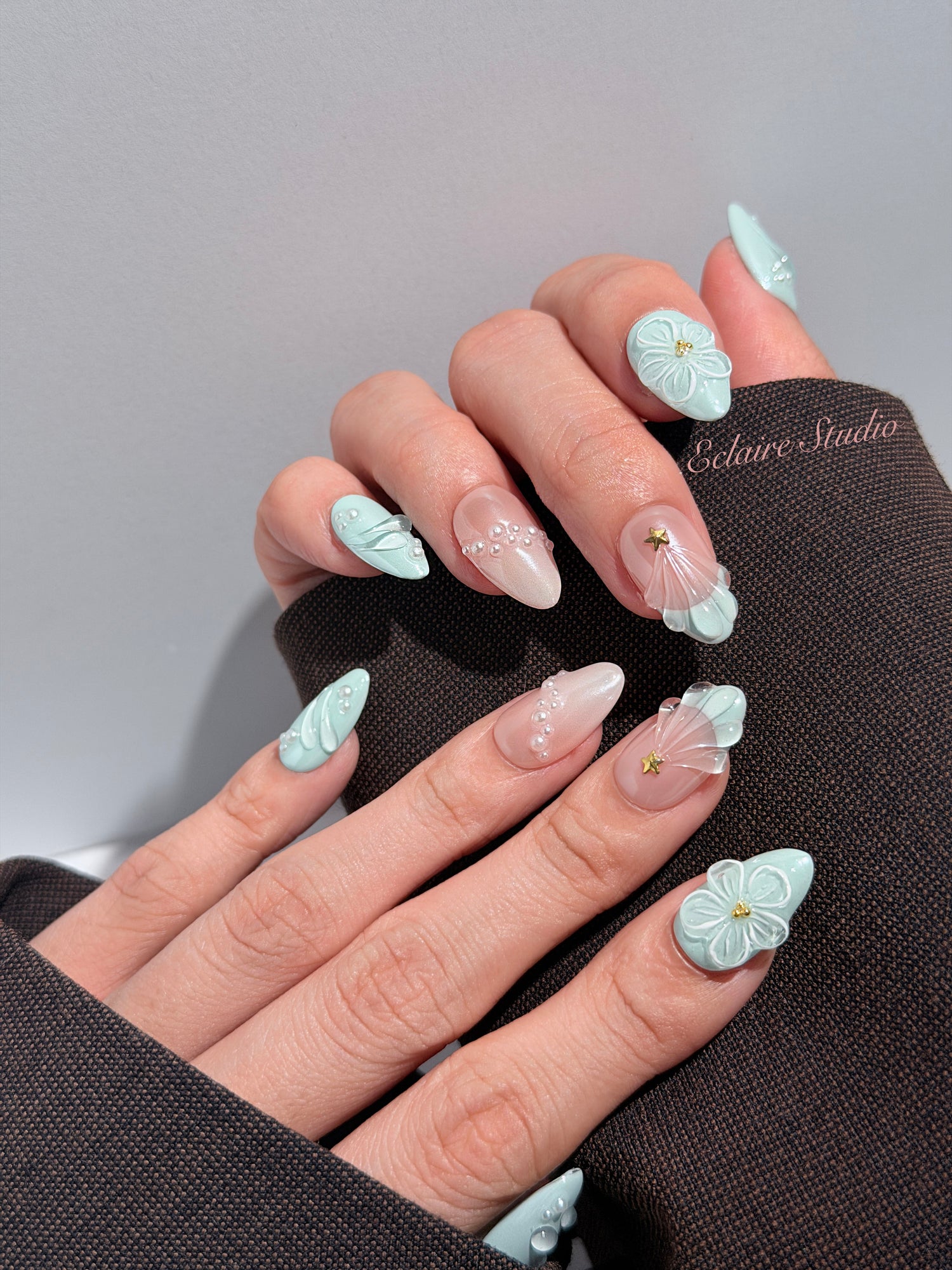 Eclaire Nail Studio 3D Summer short almond press-on nails featuring a light aqua glazed base and peachy-nude accent nails with 3D sculpted ocean waves, 3D flowers, and seashell tips with mini pearls and gold stars.