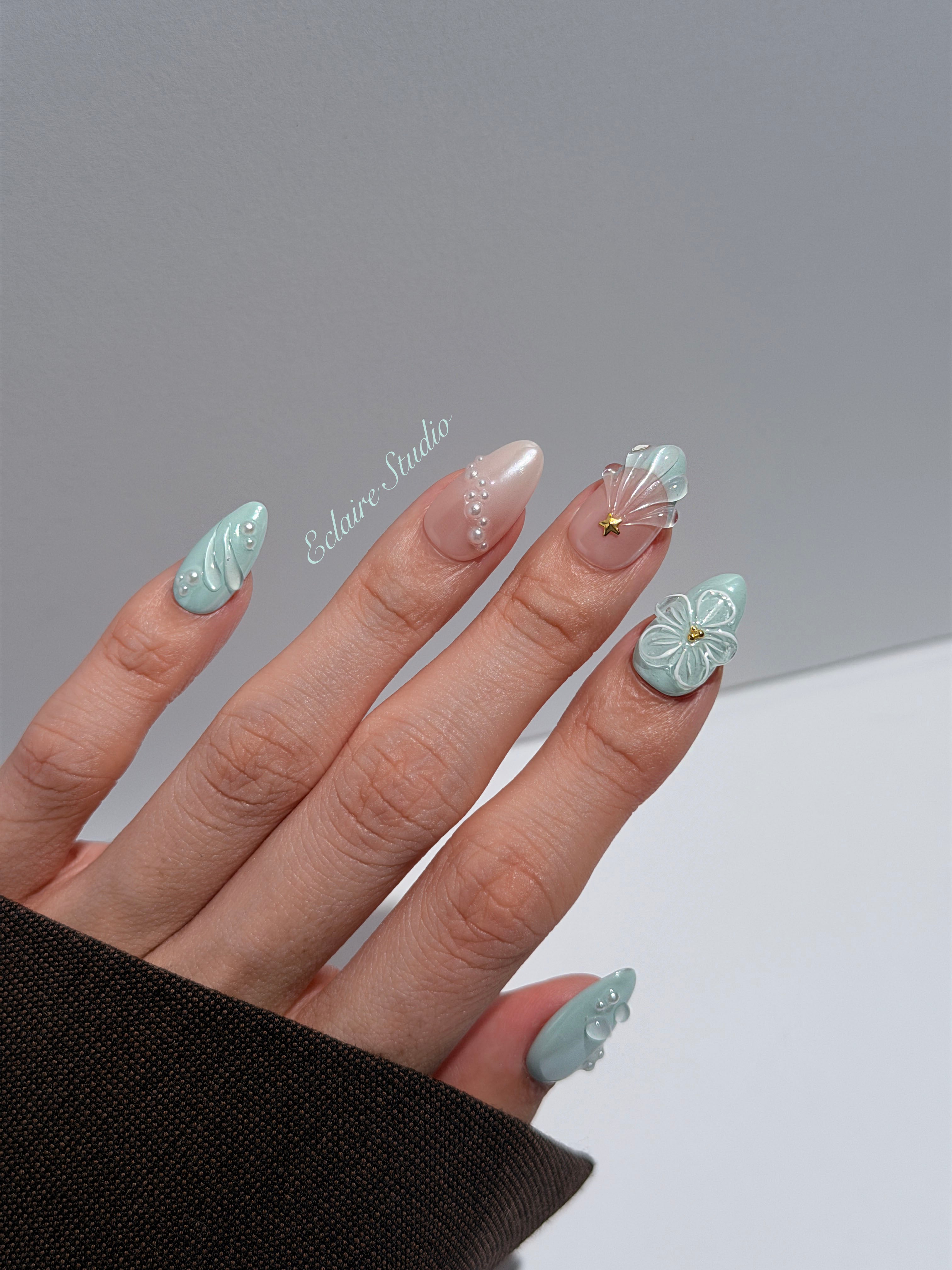 Eclaire Nail Studio 3D Summer short almond press-on nails featuring a light aqua glazed base and peachy-nude accent nails with 3D sculpted ocean waves, 3D flowers, and seashell tips with mini pearls and gold stars.