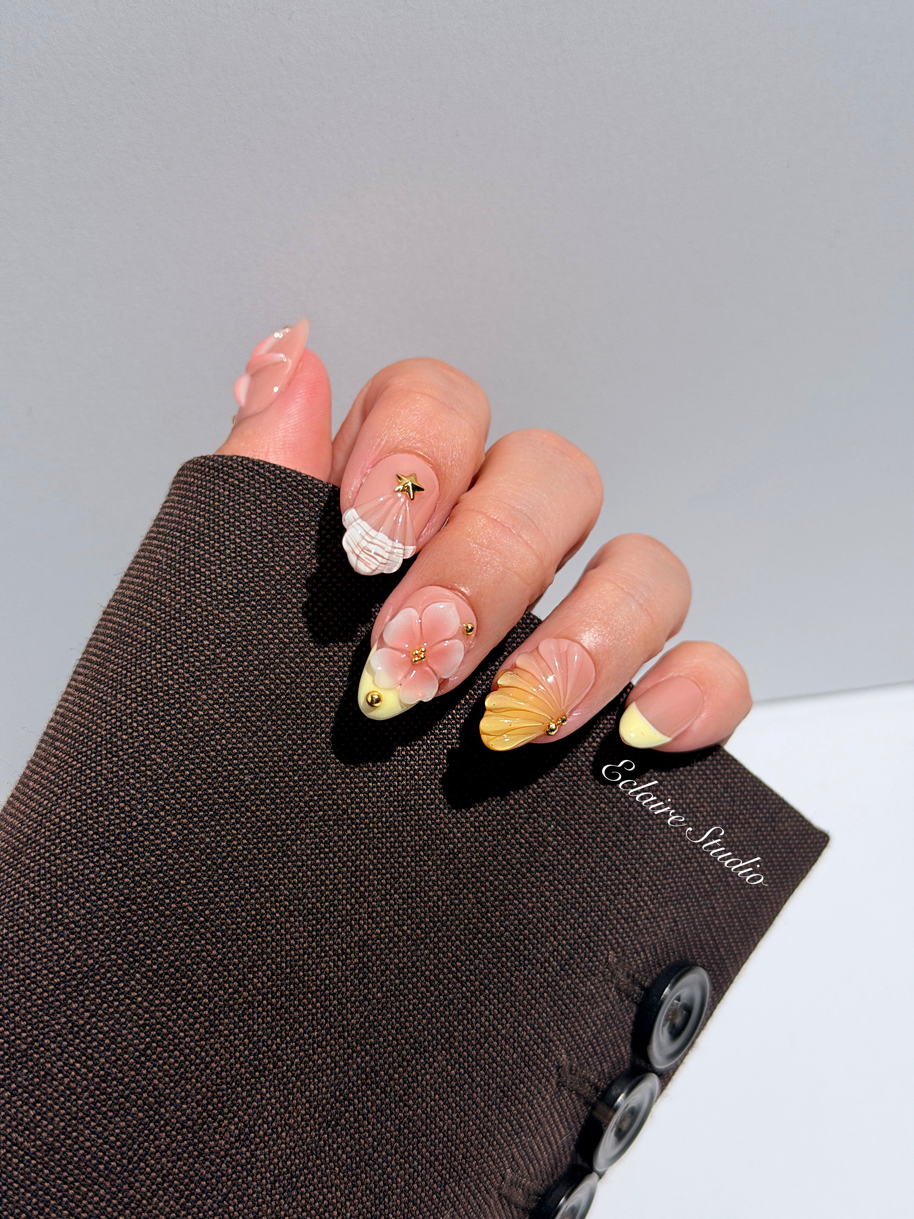 Summer Eclaire Nail Studio Luxury short almond press-on nails featuring a sheer nude base with butter yellow French tips, hand-sculpted 3D pink starfish, amber-tinted 3D seashells, and flowers with gold accents.