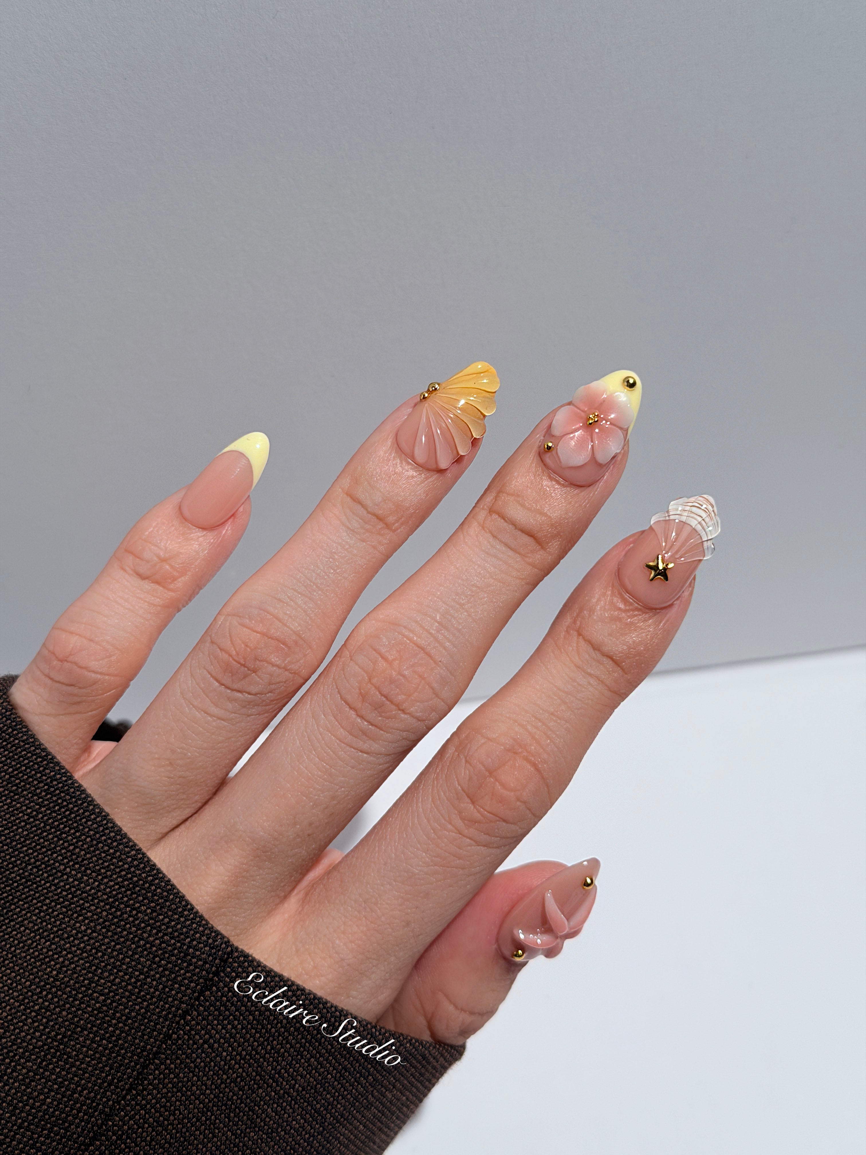 Summer Eclaire Nail Studio Luxury short almond press-on nails featuring a sheer nude base with butter yellow French tips, hand-sculpted 3D pink starfish, amber-tinted 3D seashells, and flowers with gold accents.