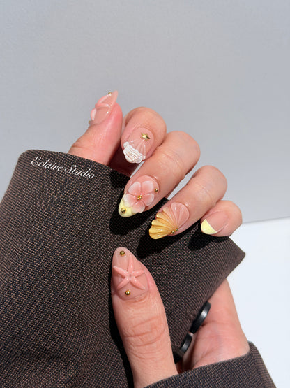 Summer Eclaire Nail Studio Luxury short almond press-on nails featuring a sheer nude base with butter yellow French tips, hand-sculpted 3D pink starfish, amber-tinted 3D seashells, and flowers with gold accents.
